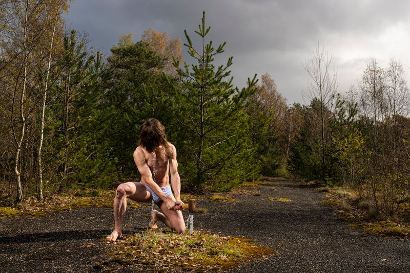 A conceptual photograph titled DRIVEN by ARJAN SPANNENBURG showing a kneeling scarred man with a sledgehammer chaining himself to the asphalt in a desolate forest landscape.