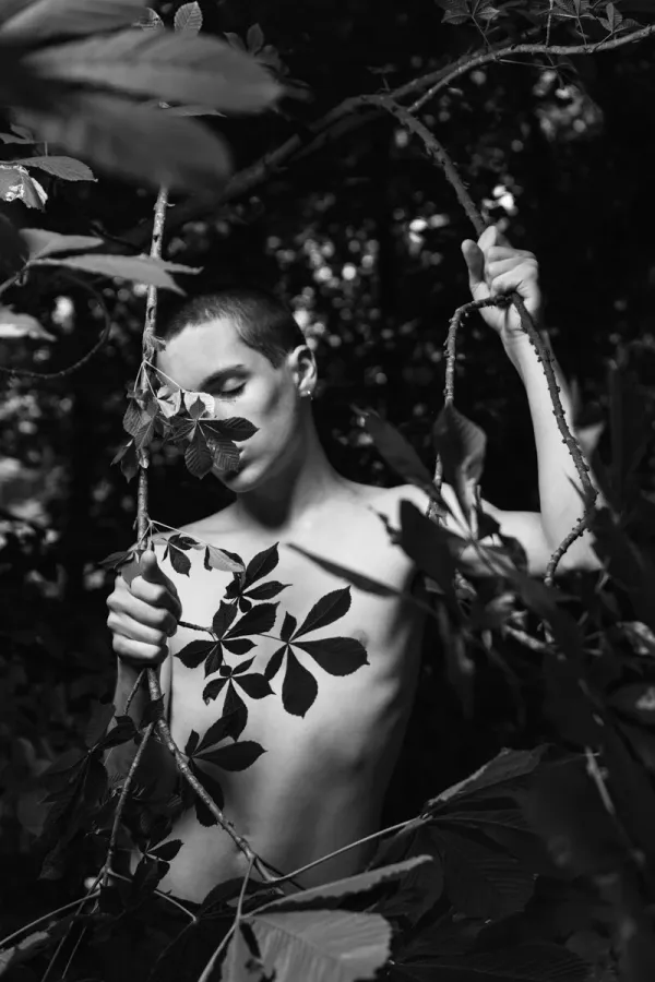 High-contrast monochrome photography of a young man with closed eyes, surrounded by chestnut tree branches and leaves, exploring themes of truth and identity in ARJAN SPANNENBURG'S ESTRANGED series.