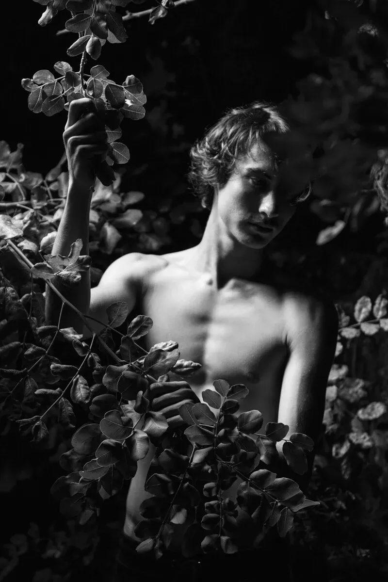 High-contrast monochrome photography of a young man with wavy hair partially hidden by locust tree leaves, exploring themes of home and understanding in ARJAN SPANNENBURG'S ESTRANGED series.