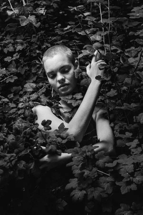 A black and white conceptual fine art portrait of ROBIN for the ESTRANGED series by ARJAN SPANNENBURG, featuring a person with a shaved head among dense leaves.