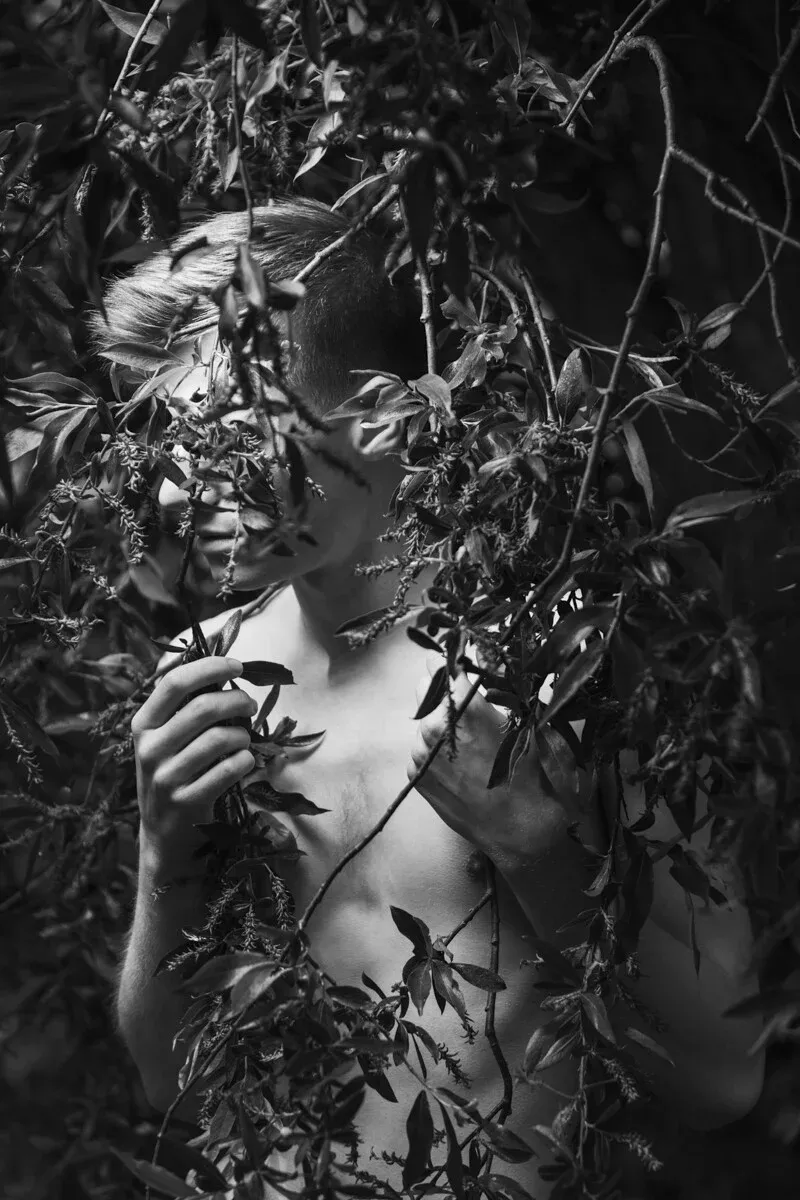 A black and white conceptual portrait of YANNICK for the ESTRANGED series by ARJAN SPANNENBURG, showing a man partially hidden by pine branches.